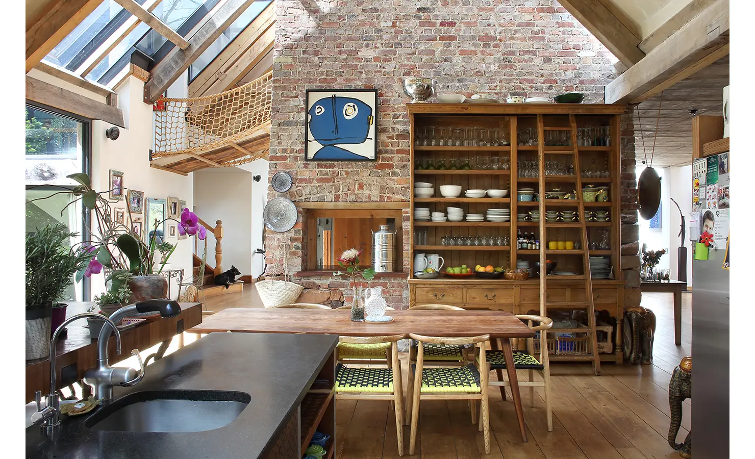 Perfect Hideaways. Spacious rustic kitchen with brick walls and wooden beams, featuring an open shelving unit stocked with dishes and glassware, a long wooden dining table, and a countertop with a sink. Natural light pours in through large skylights, highlighting the eclectic decor and vibrant plants. Gaia, Constantia.