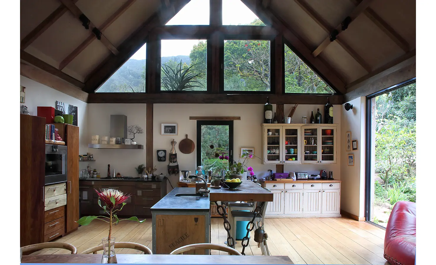 Perfect Hideaways. Rustic kitchen interior with high ceilings and large windows offering a view of lush greenery. Features a wooden island, modern appliances, and open shelving decorated with plants and colorful ceramics. Natural light enhances the cozy ambiance. Gaia, Constantia.