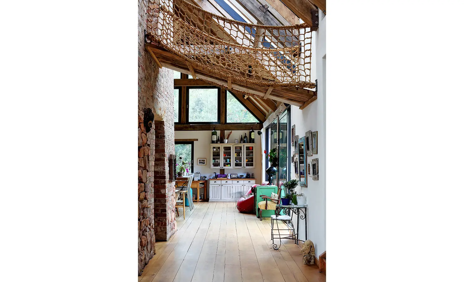 Perfect Hideaways. A rustic living area with wooden beams, stone walls, and a netted loft. Large windows provide natural light. Decor includes plants, artwork, and various furniture pieces. Gaia, Constantia.