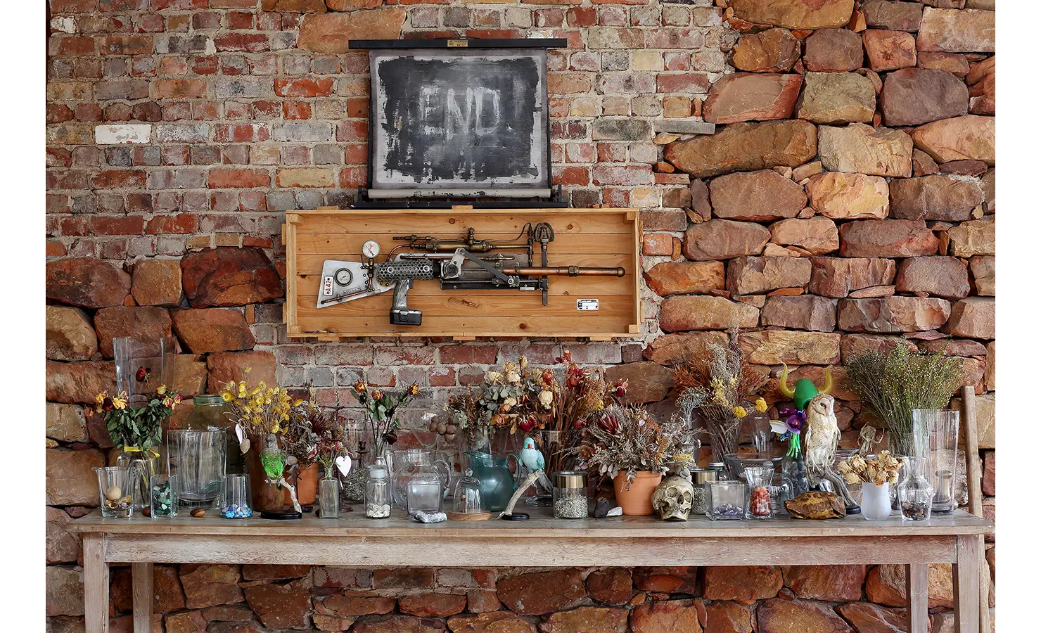Perfect Hideaways. A rustic brick and stone wall displays a chalkboard with END written on it, a steampunk-style gun, and a table below featuring an assortment of dried flowers and glass jars. Gaia, Constantia.