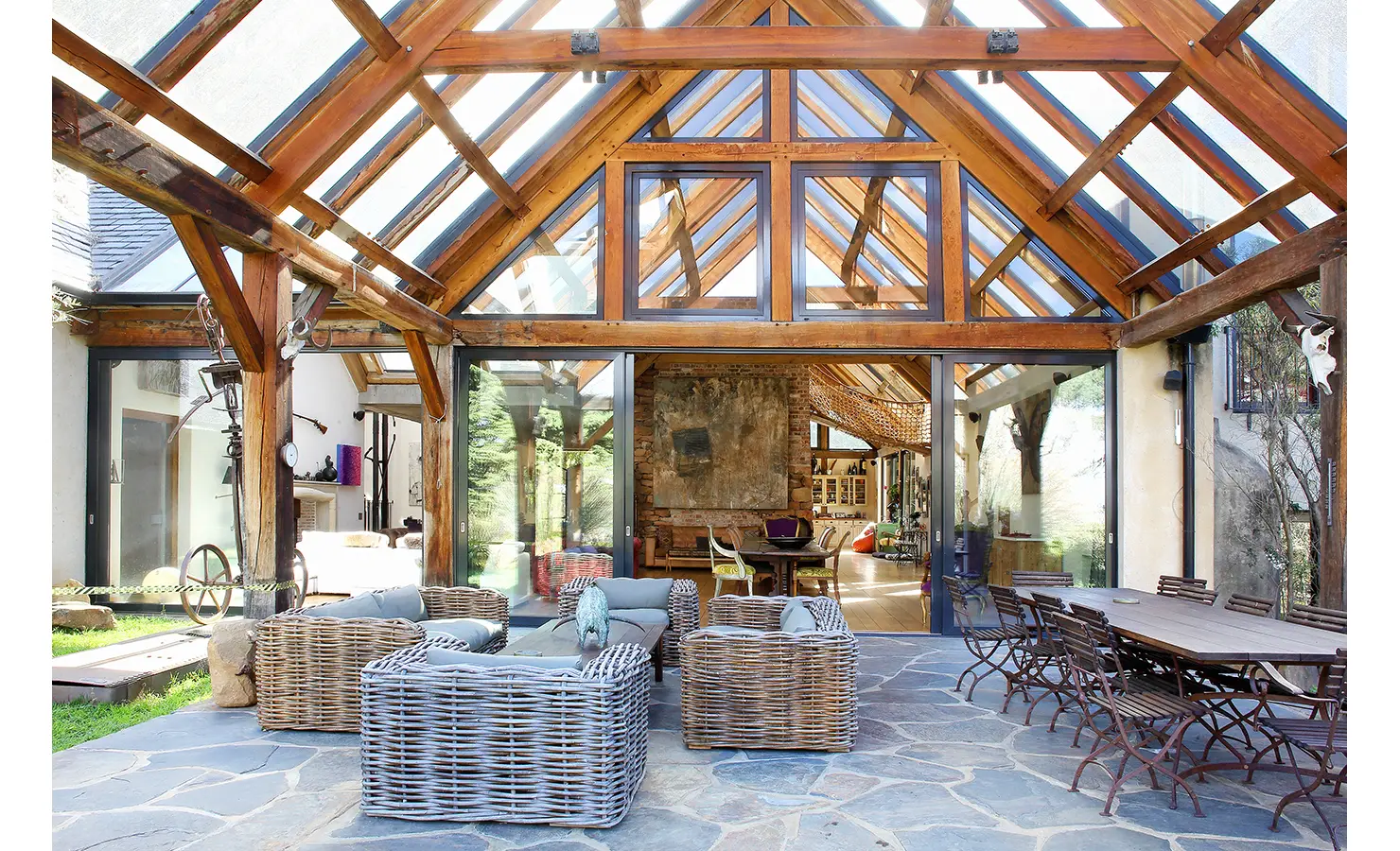 Perfect Hideaways. A spacious, rustic conservatory with a glass roof and wooden beams. Furnished with wicker sofas, a large dining table, and chairs on a stone tile floor. The interior features a mix of wood and natural light. Gaia, Constantia.