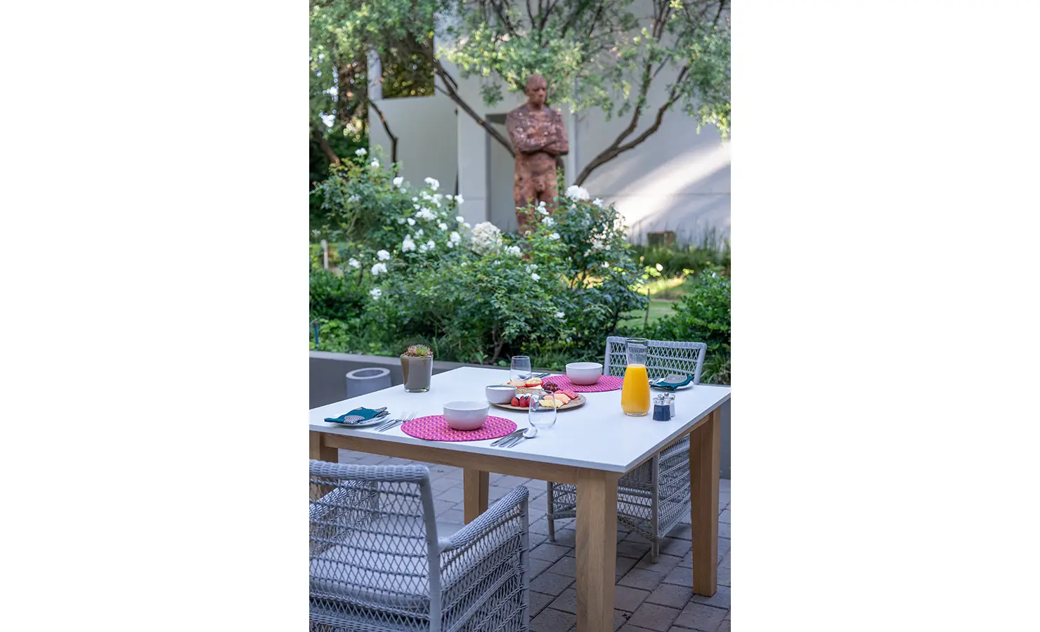 Perfect Hideaways: A tranquil outdoor breakfast table is set in a lush garden courtyard, framed by flowering greenery and a striking sculpture. AtholPlace Hotel & Villa, Atholl.