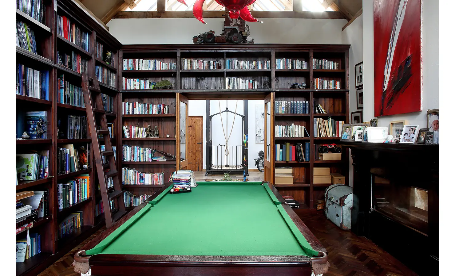 Perfect Hideaways. A cozy library with wooden bookshelves filled with books, surrounding a green pool table in the center. A ladder leans against a shelf, and a red sculpture hangs from the ceiling. Photos and decorative items are displayed on a mantelpiece. Gaia, Constantia.