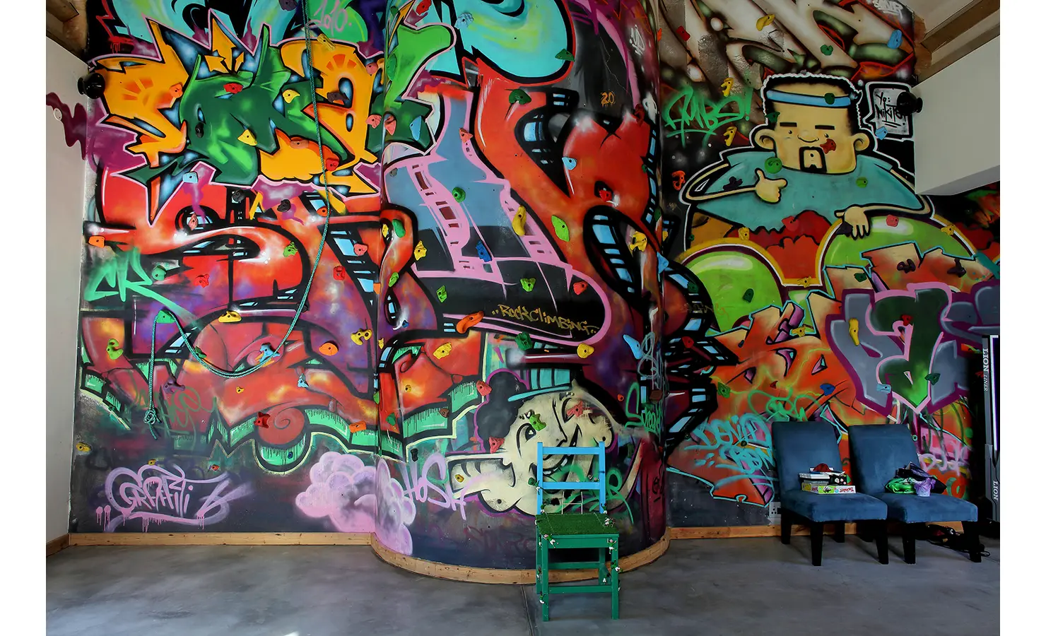 Perfect Hideaways. A vibrant wall covered in colorful graffiti art features a variety of abstract shapes, characters, and lettering. In front of the wall, there are two chairs: one green, one dark gray. The floor is concrete, adding an industrial feel. Gaia, Constantia.