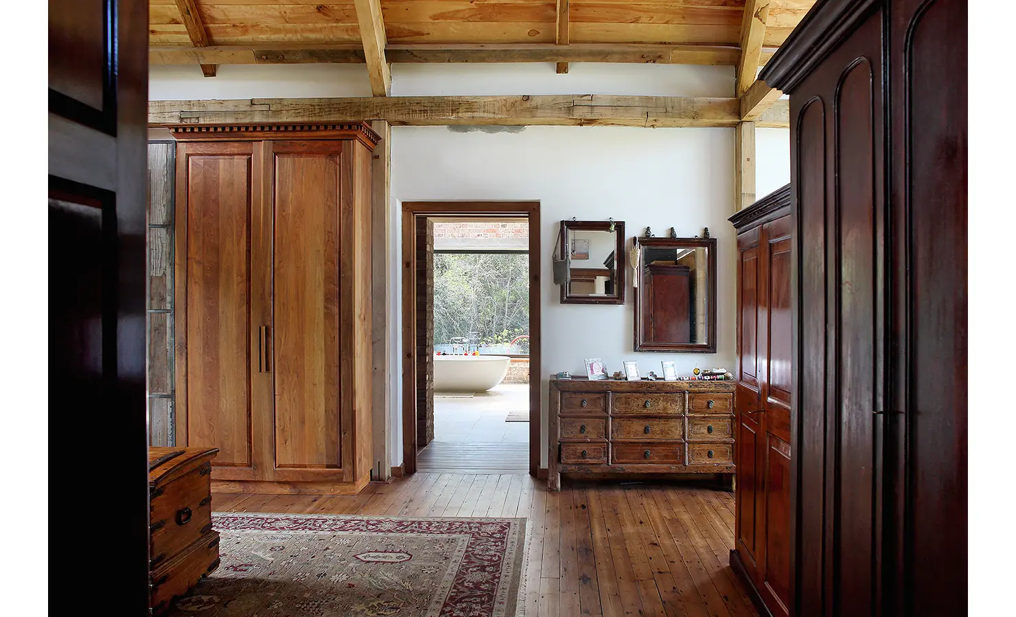 Perfect Hideaways. A rustic bedroom with wooden furniture, including a wardrobe and dresser. The wooden floor is partially covered by a rug. An open doorway reveals a bathroom with a freestanding tub by a large window overlooking a lush landscape. Gaia, Constantia.
