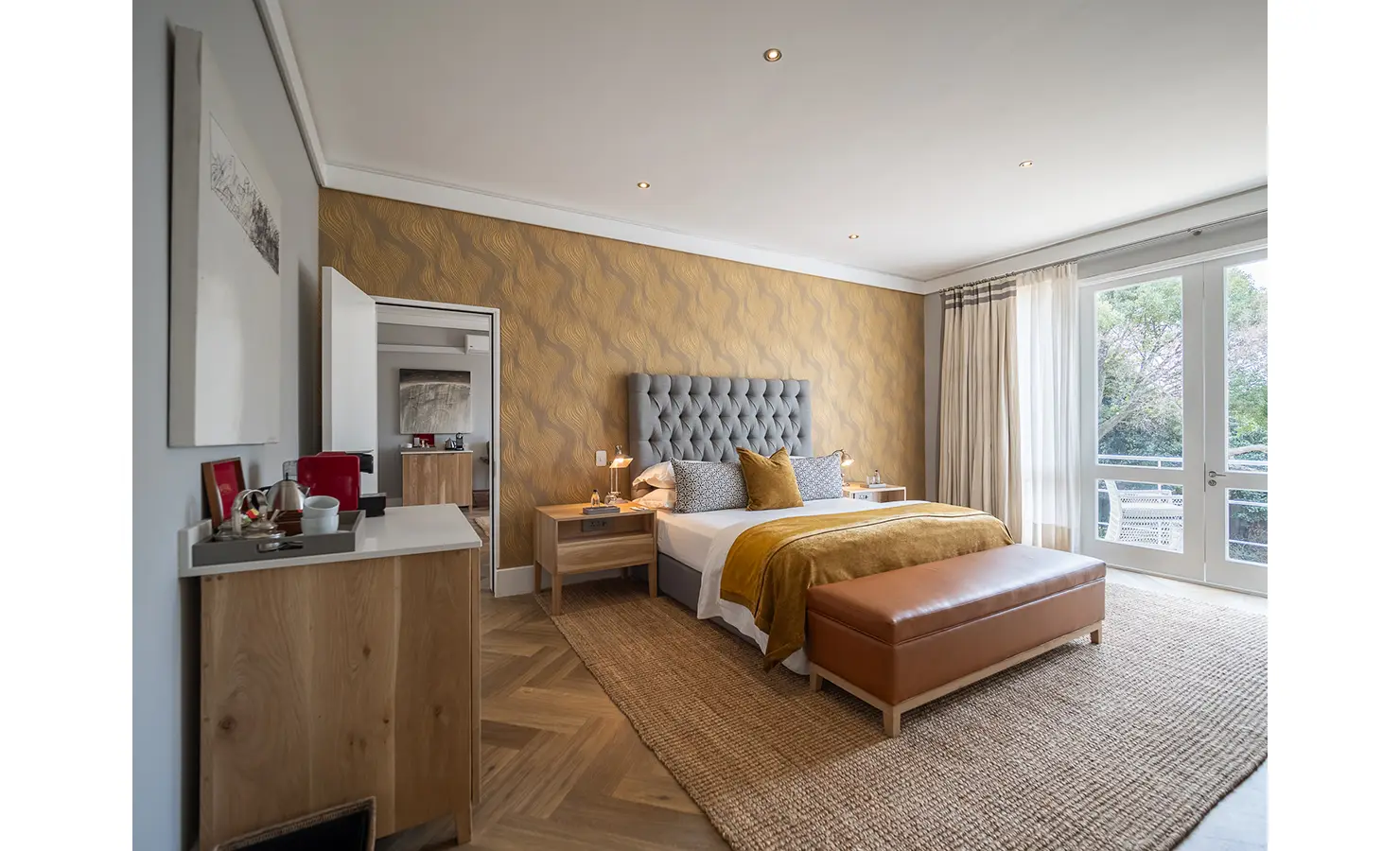 Perfect Hideaways: A warm-toned bedroom with tufted grey headboard, amber throws, and natural textures opens onto a balcony with leafy views. AtholPlace Hotel & Villa, Atholl.