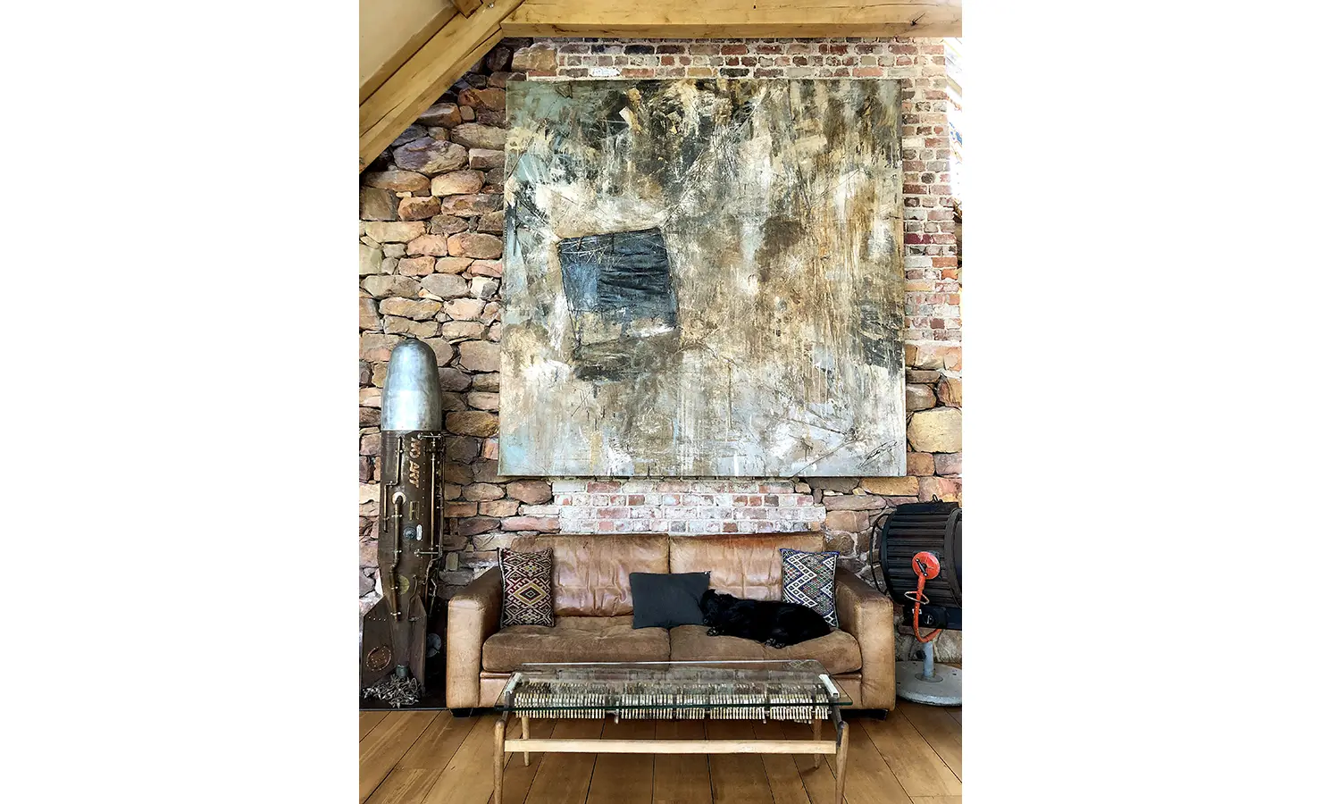 Perfect Hideaways. A rustic living room features a large abstract painting on a brick-and-stone wall. A brown leather sofa below has patterned cushions and a black cat lounging. A vintage rocket and industrial fan add eclectic touches. A woven coffee table is in front. Gaia, Constantia.