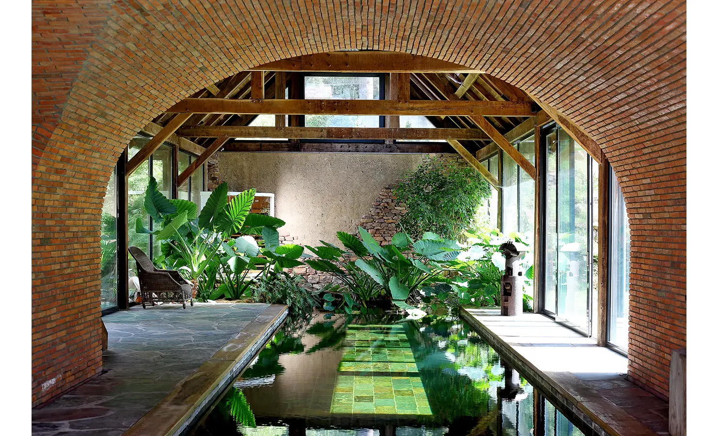 Perfect Hideaways. A serene indoor pool surrounded by lush green plants under a rustic wooden and brick arched ceiling. Natural light streams in through large windows on both sides, illuminating the stone walkway and creating reflections on the water. Gaia, Constantia.