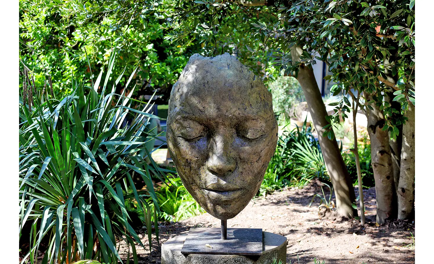 Perfect Hideaways. A large bronze sculpture of a peaceful face rests on a pedestal surrounded by lush green foliage and trees in a garden setting. The sculpture features closed eyes and a serene expression. Gaia, Constantia.