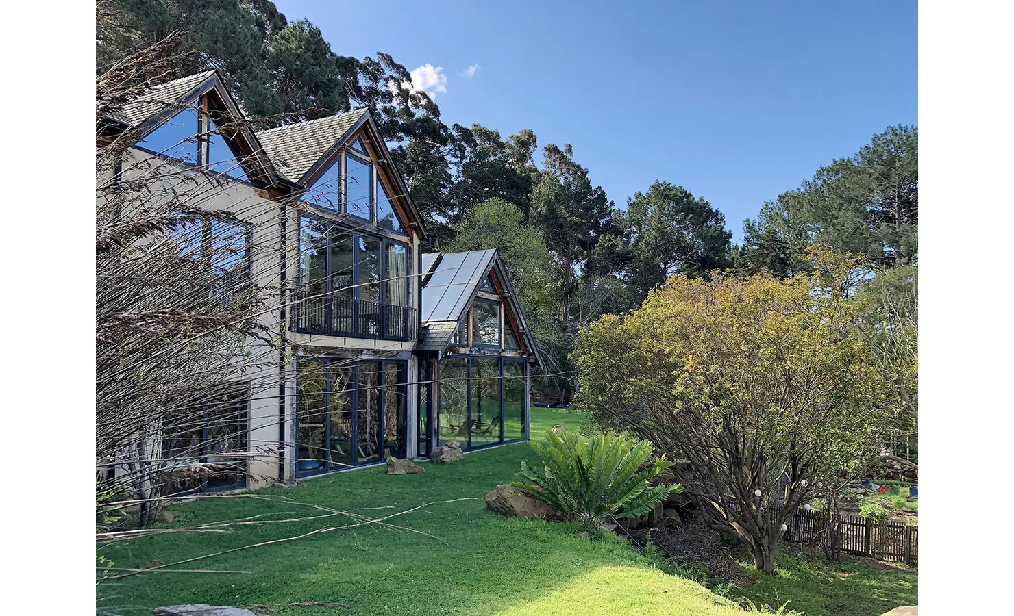 Perfect Hideaways. A modern house with large glass windows sits amidst lush greenery and tall trees under a clear blue sky. The building features sloped roofs and is surrounded by a well-maintained lawn and garden. Gaia, Constantia.