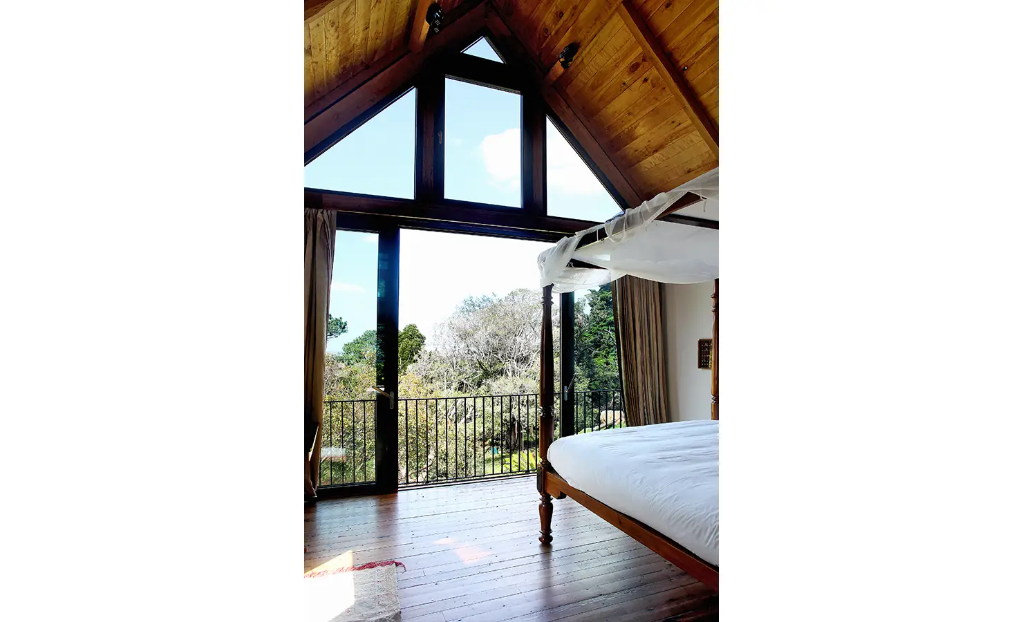 Perfect Hideaways. A cozy bedroom with a wooden canopy bed and a large triangular window offering a view of trees and blue sky. The room features wooden floors, a high ceiling, and natural light streaming in through the glass doors leading to a balcony. Gaia, Constantia.
