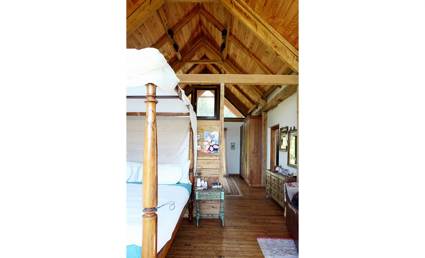 Perfect Hideaways. A rustic bedroom with a high wooden ceiling and exposed beams. It features a wooden four-poster bed with white bedding, a small vintage table, a dresser with a mirror, and artwork on the wall. The room has wooden floors and natural light. Gaia, Constantia.