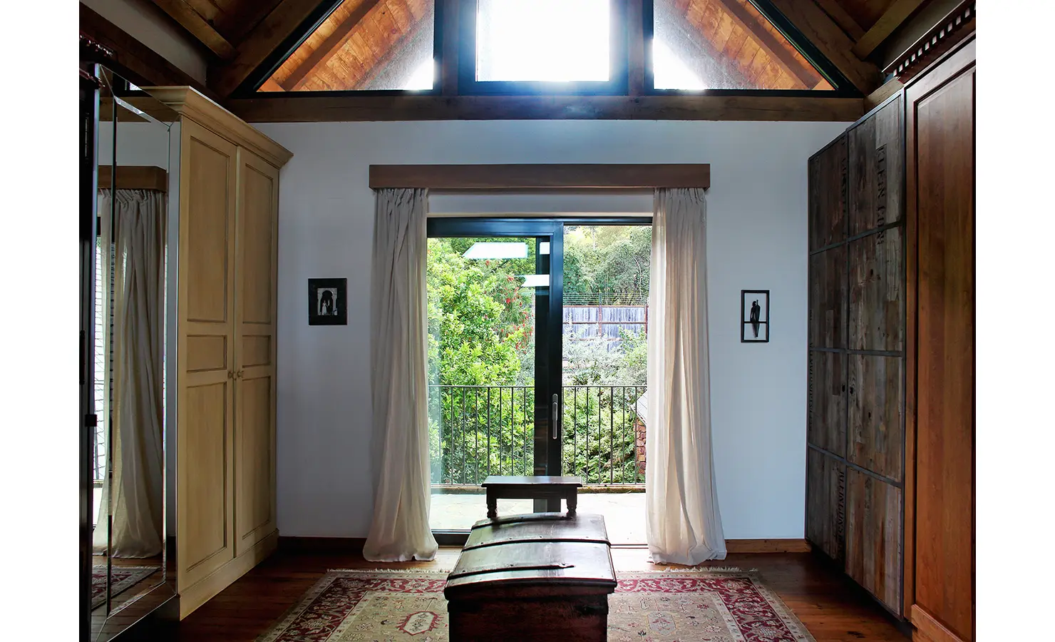 Perfect Hideaways. A cozy room with a high wooden ceiling, featuring a large glass door opening to a balcony with greenery. It has wooden wardrobes, a trunk-like table, and a patterned rug, with sunlight filtering through a triangular window above the door. Gaia, Constantia.