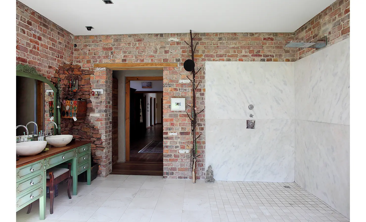Perfect Hideaways. A rustic bathroom featuring brick walls and a marble shower area. A green vanity holds dual sinks with round mirrors. A hallway is visible through an open wooden door. Decorative items include a wall-mounted plant and a small statue. Gaia, Constantia.