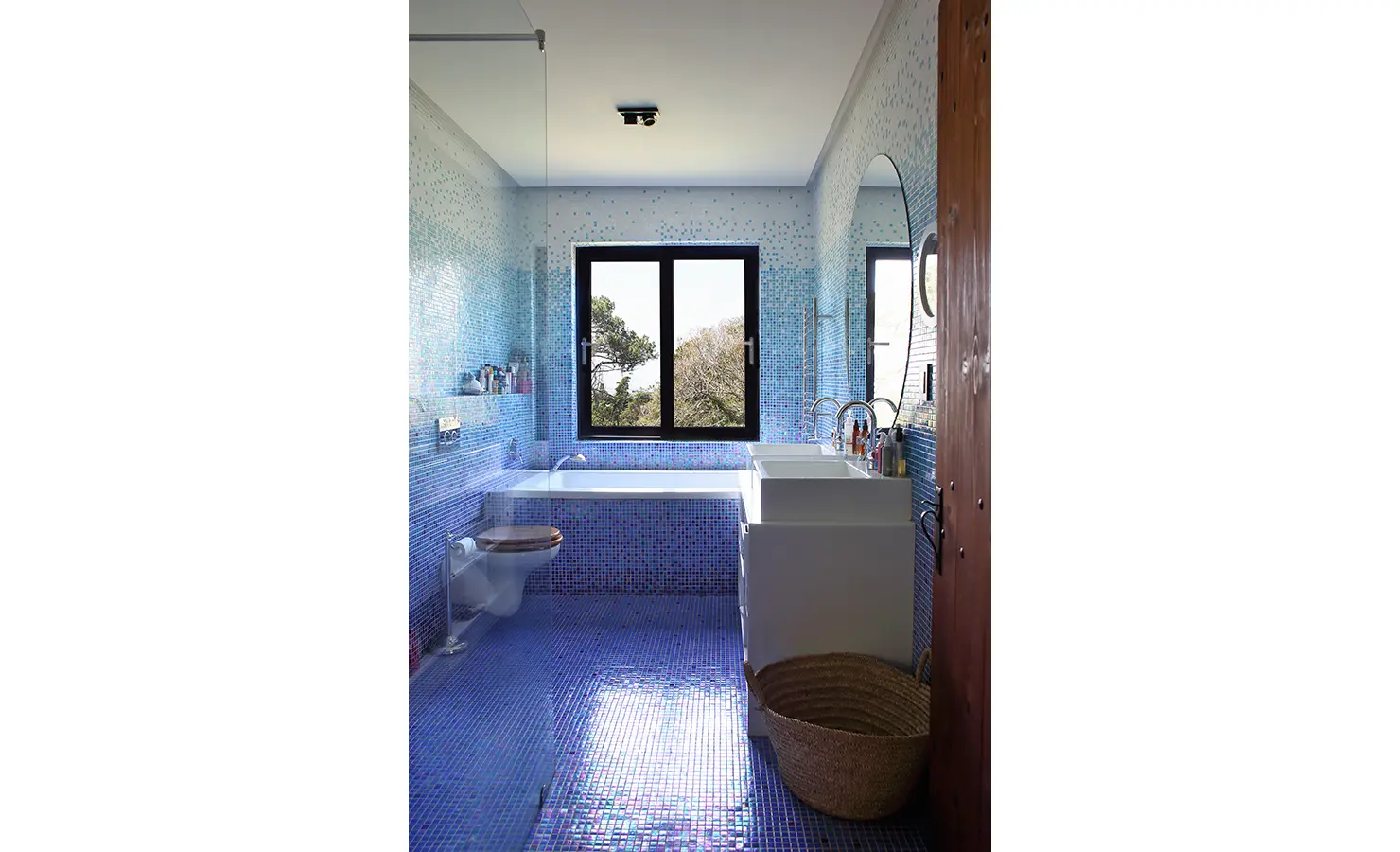 Perfect Hideaways. A modern bathroom with blue mosaic tiles covering the walls and floor. It features a large window with a view of trees, a white bathtub, a glass shower enclosure, a white sink, and a wooden door. A woven basket sits on the floor. Gaia, Constantia.