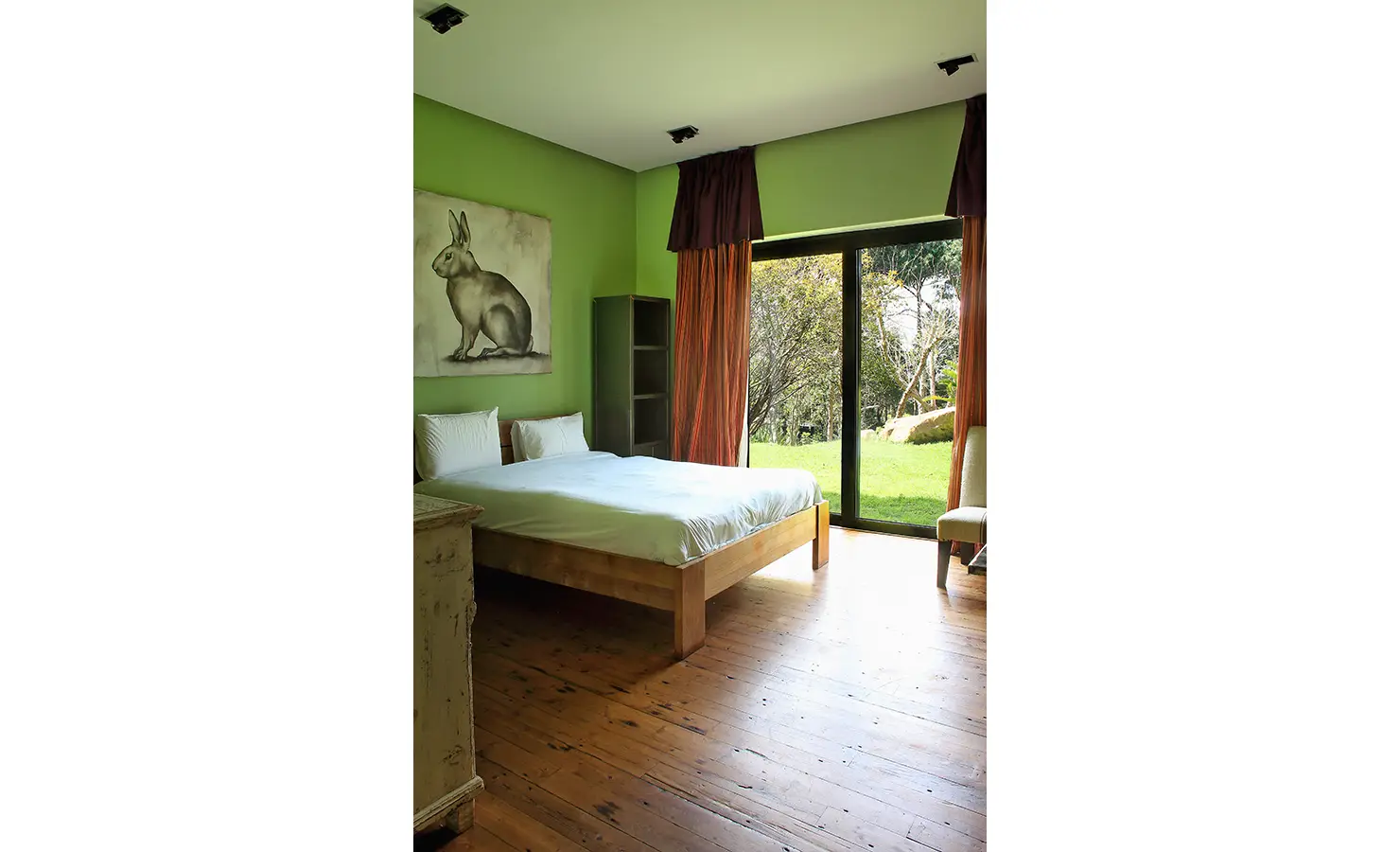 Perfect Hideaways. A bedroom with green walls features a wooden bed with white linens, an animal painting above the headboard, and a rustic chest. Large glass doors with red curtains open to a grassy outdoor area, offering a view of trees and rocks. Gaia, Constantia.