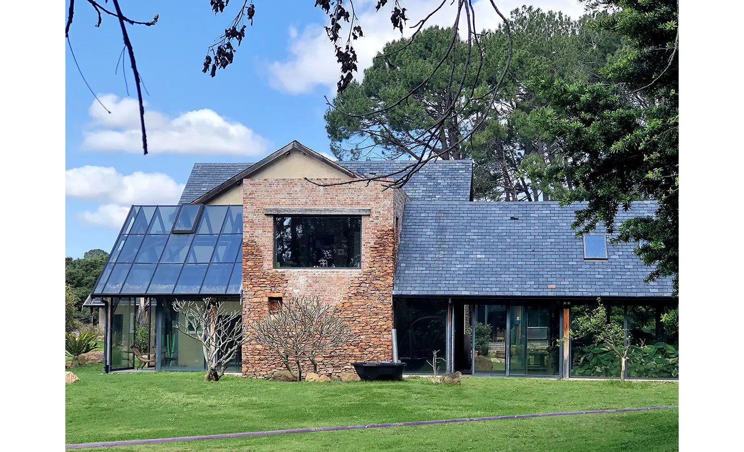 Perfect Hideaways. A modern house with a mix of glass and brick walls, featuring a large glass facade and a central brick chimney. Surrounded by lush green grass and trees, the sky is clear with a few scattered clouds. Gaia, Constantia.