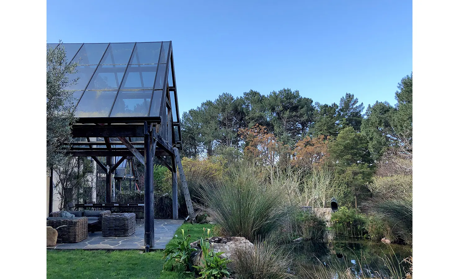 Perfect Hideaways. An outdoor seating area with wicker furniture is sheltered by a large glass canopy. The scene is surrounded by lush greenery and tall trees under a clear blue sky, creating a serene, natural atmosphere. Gaia, Constantia.