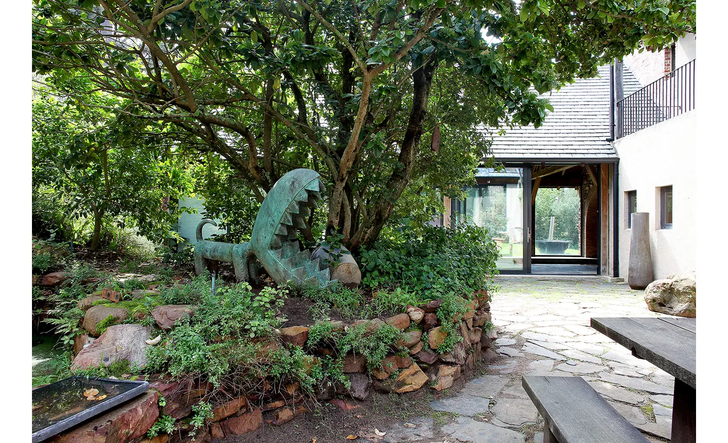 Perfect Hideaways. A lush garden with a large, green, whimsical sculpture resembling a plant with jagged leaves sits among bushes and trees. The scene is set beside a stone path leading to a modern house with large glass windows and wooden accents. Gaia, Constantia.