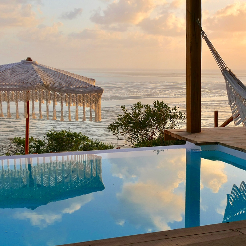 Perfect Hideaways – A serene oceanfront scene at sunrise featuring a still turquoise pool reflecting the sky, a crochet parasol, and a white woven hammock hanging from a wooden deck overlooking expansive tidal flats glowing in warm golden light. Azulik Villa, The Sanctuary Private Reserve