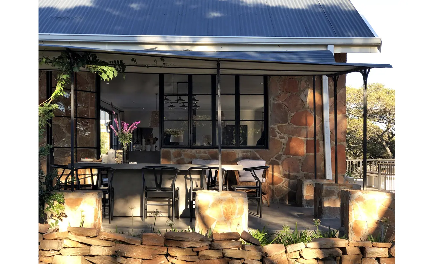 Perfect Hideaways: A charming patio area with stone walls, black-framed windows, and a bar counter surrounded by natural stone landscaping and shaded greenery. Owlswood, Buffelspoort.
