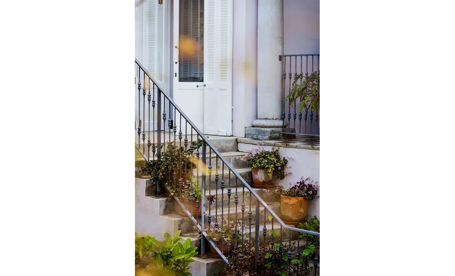 Perfect Hideaways: A delicate staircase framed by wrought iron railings, terracotta pots, and lush greenery. The soft pastel hues of the façade evoke a romantic, European-style courtyard. Dorp Hotel, Bo Kaap.