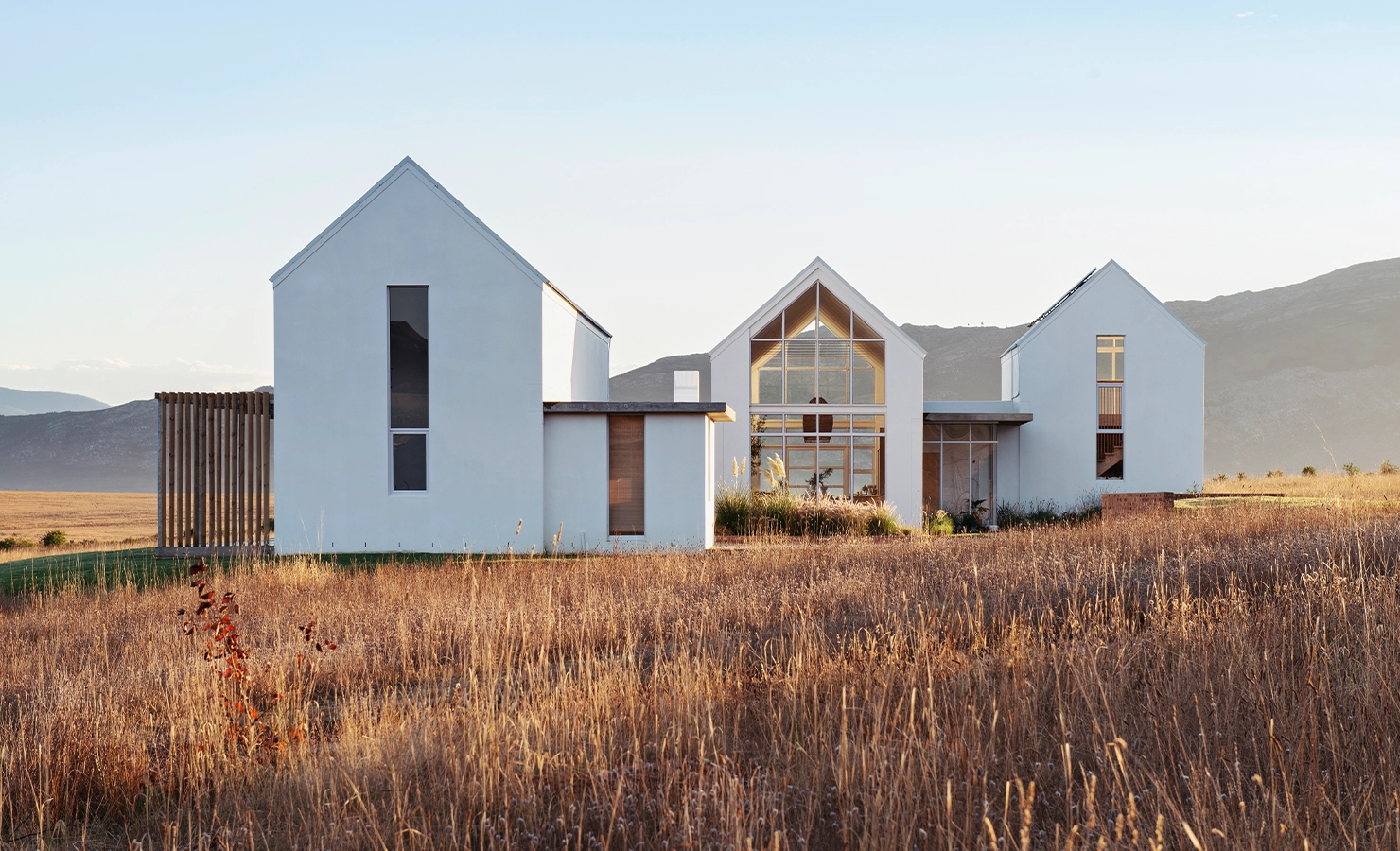 Perfect Hideaways: Contemporary white-gabled farmhouse set in golden grasslands with mountain views, featuring expansive glass windows and minimalist architectural lines, Thuúla Haven, Hemel-en-Aarde.