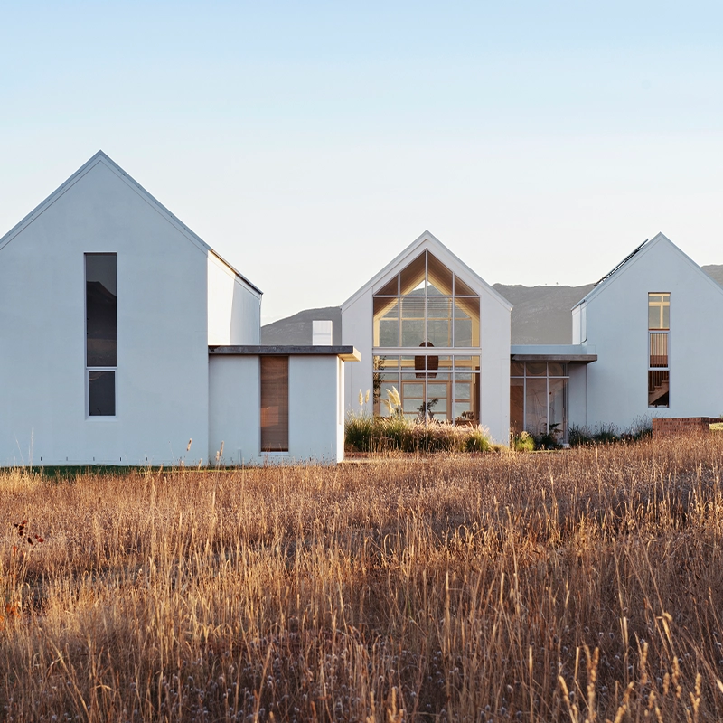 Perfect Hideaways: Contemporary white-gabled farmhouse set in golden grasslands with mountain views, featuring expansive glass windows and minimalist architectural lines, Thuúla Haven, Hemel-en-Aarde.