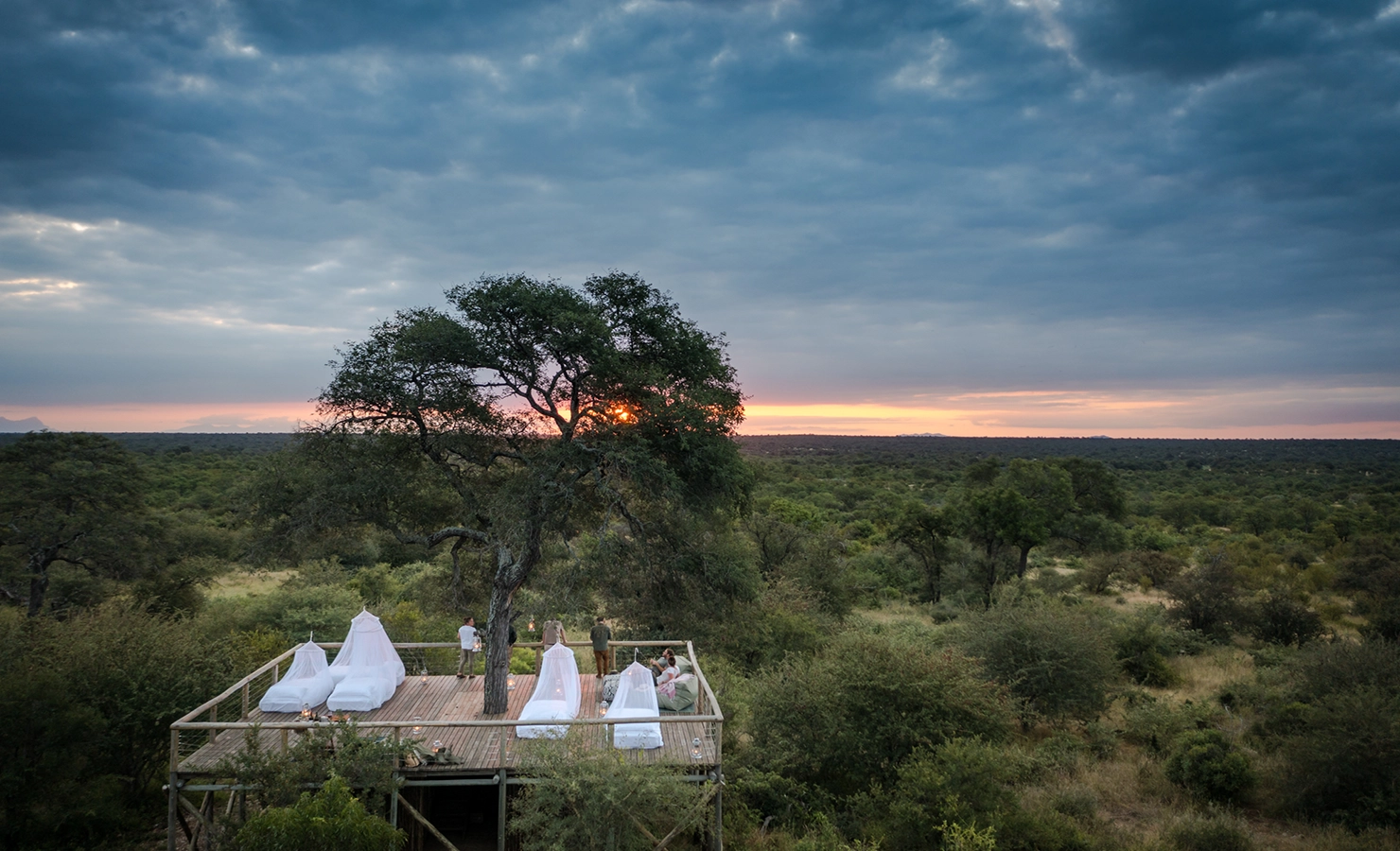 Perfect Hideaways: A panoramic view of a raised deck nestled in the bush, featuring outdoor beds and mosquito nets, with a spectacular sunset over the wilderness. Tulela Lodge, Klaserie Private Nature Reserve.