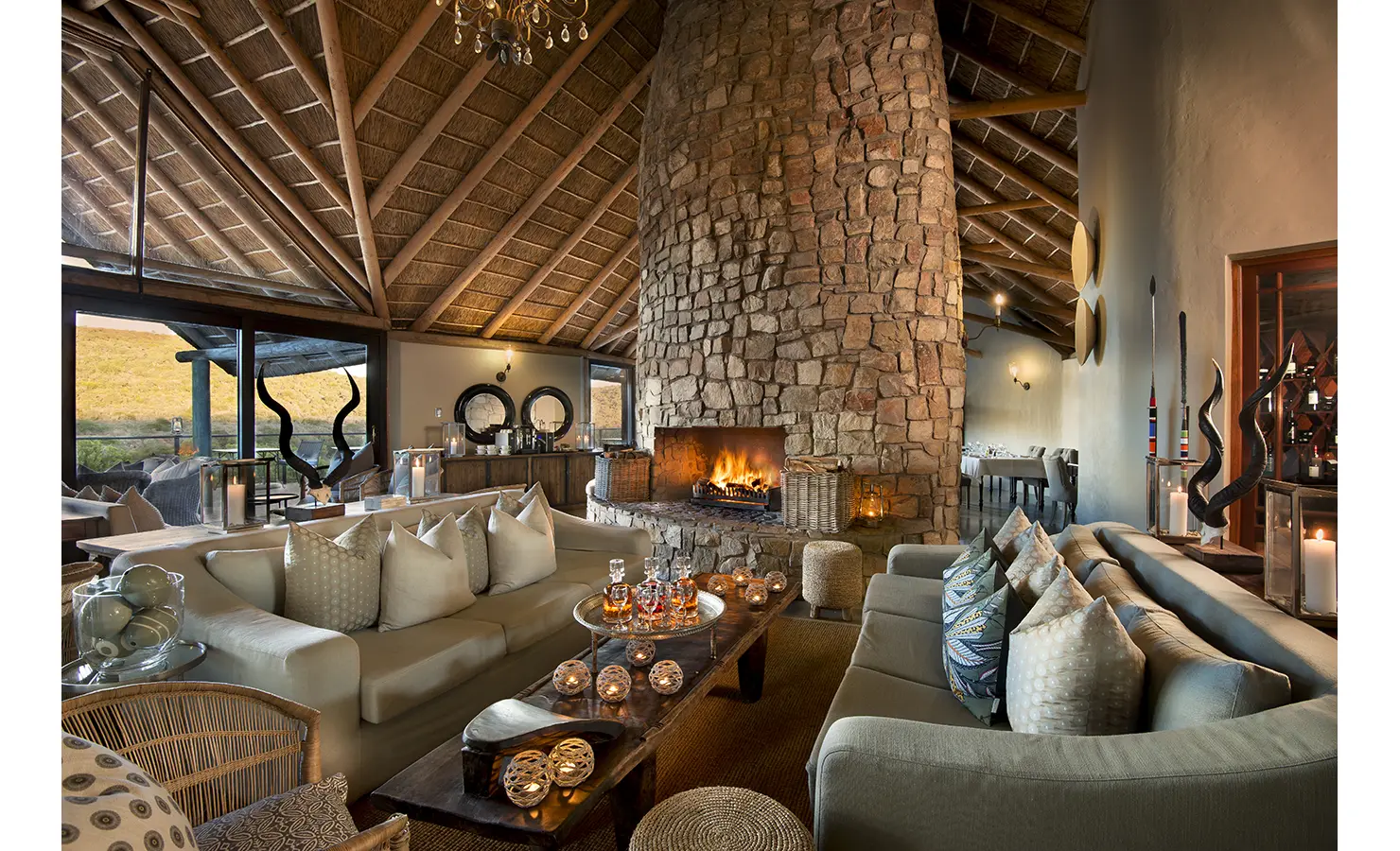 Perfect Hideaways: A beautifully styled lodge interior featuring a grand stone fireplace, plush seating, and soft lighting under a high thatched ceiling. The warm, earthy tones create a welcoming and luxurious retreat. Great Fish River Lodge, Kwandwe Private Game Reserve.