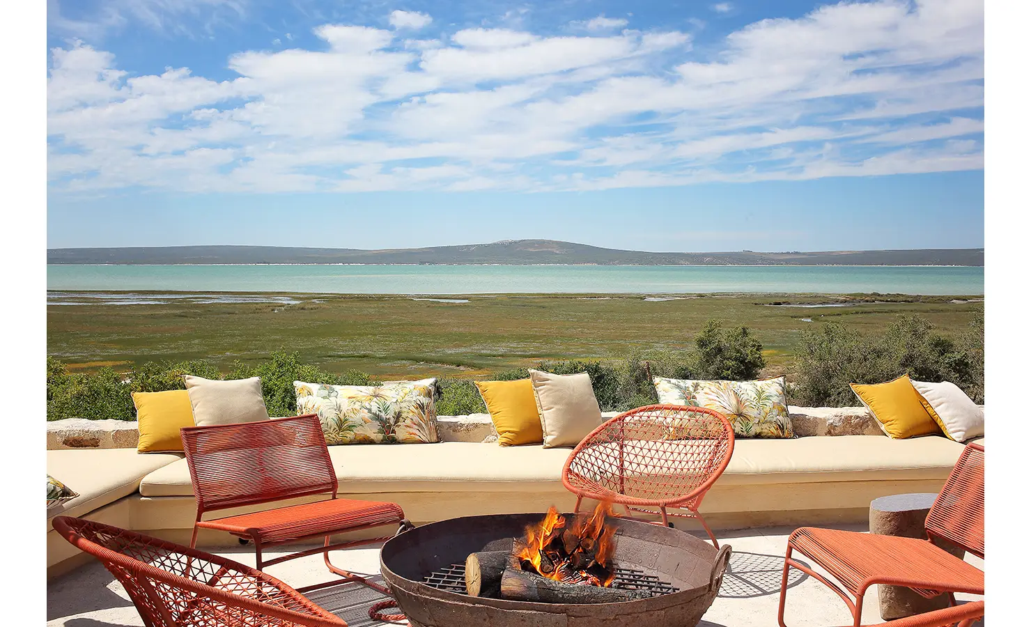 Perfect Hideaways: A stunning outdoor fire pit area with bright orange chairs and a long built-in bench adorned with yellow and patterned cushions, overlooking a vast stretch of green marshland and tranquil turquoise waters under a blue sky. Flamingo, Churchhaven.