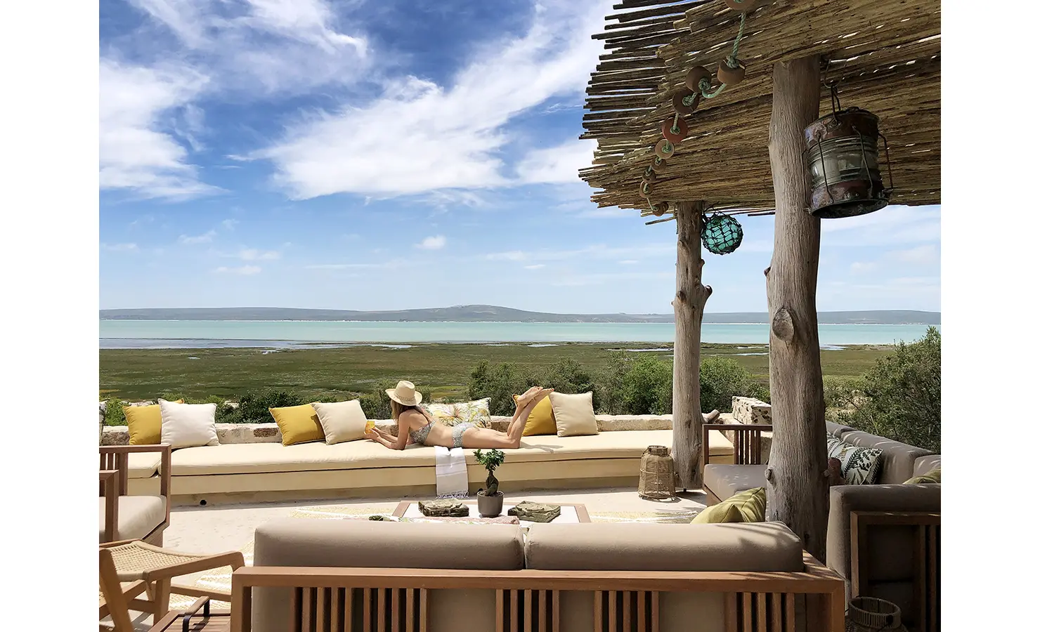 Perfect Hideaways: A serene outdoor lounge space featuring a wooden pergola, a long cushioned bench, and rustic wooden pillars, framing an uninterrupted view of the lagoon and rolling greenery under a vast sky. Flamingo, Churchhaven.