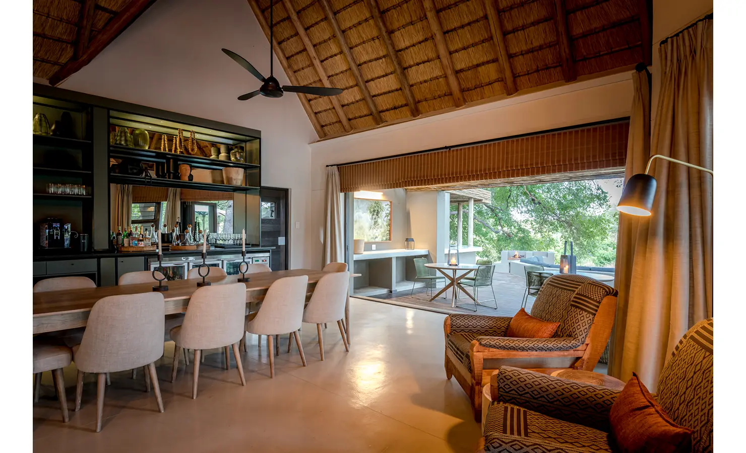 Perfect Hideaways: A modern dining area featuring a long wooden table with comfortable upholstered chairs, leading to an outdoor patio with views of the lush wilderness. Tulela Lodge, Klaserie Private Nature Reserve.