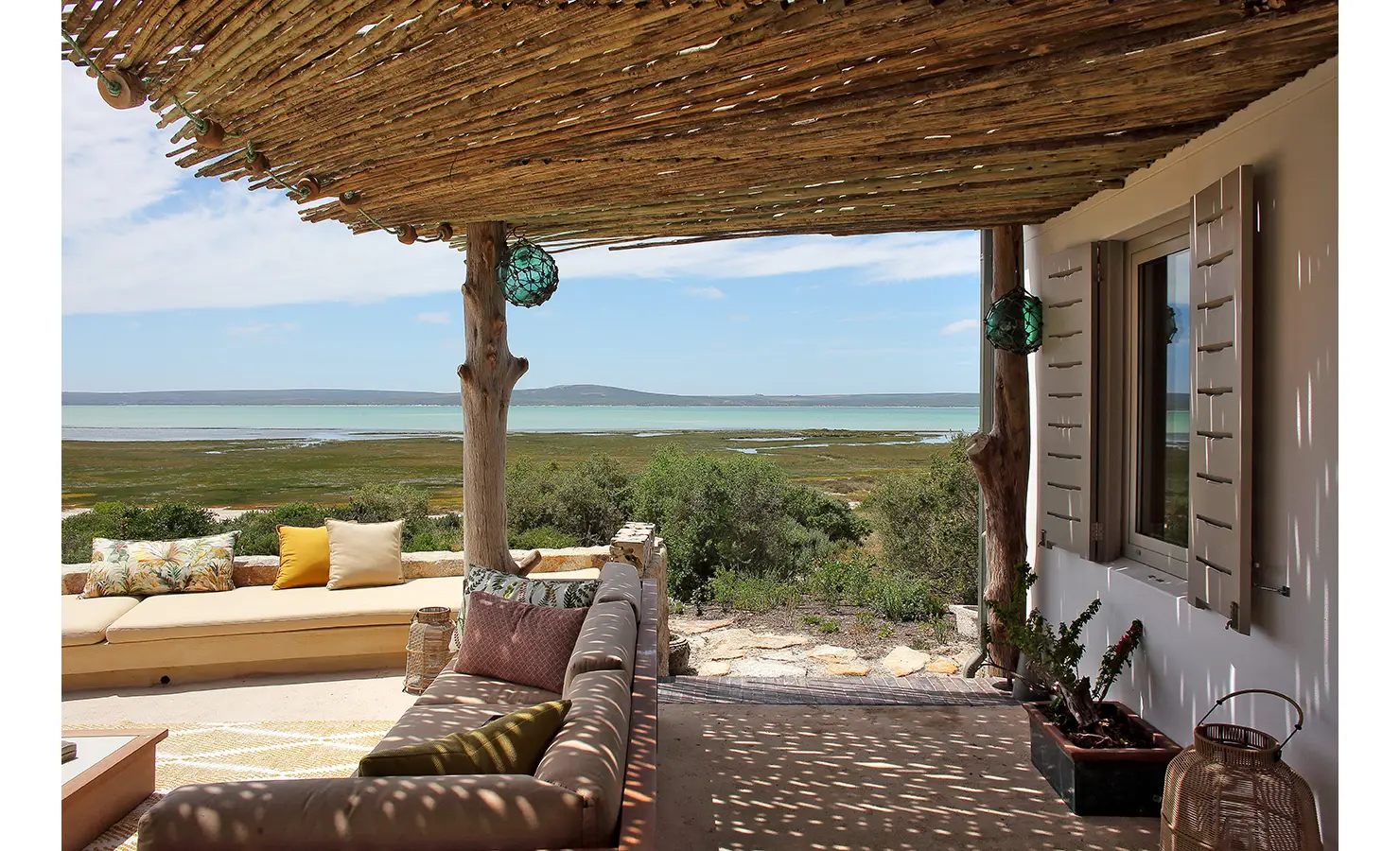 Perfect Hideaways: A guest enjoys the sun on a stylish outdoor seating area, sipping a drink while lying on a cushioned bench with yellow and neutral pillows, surrounded by the beauty of nature and the shimmering waters in the distance. Flamingo, Churchhaven.