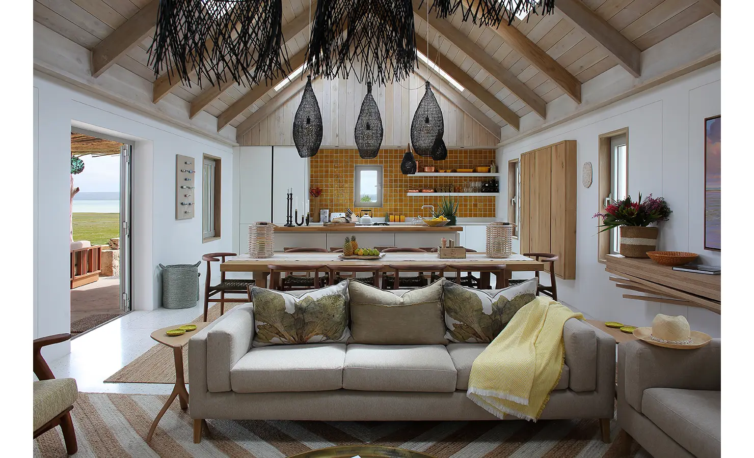 Perfect Hideaways: A spacious open-plan living and dining area with vaulted ceilings, featuring a soft-toned sofa, a rustic wooden dining table, and a stylish kitchen with mustard-hued tiles, complemented by striking woven pendant lights. Flamingo, Churchhaven.