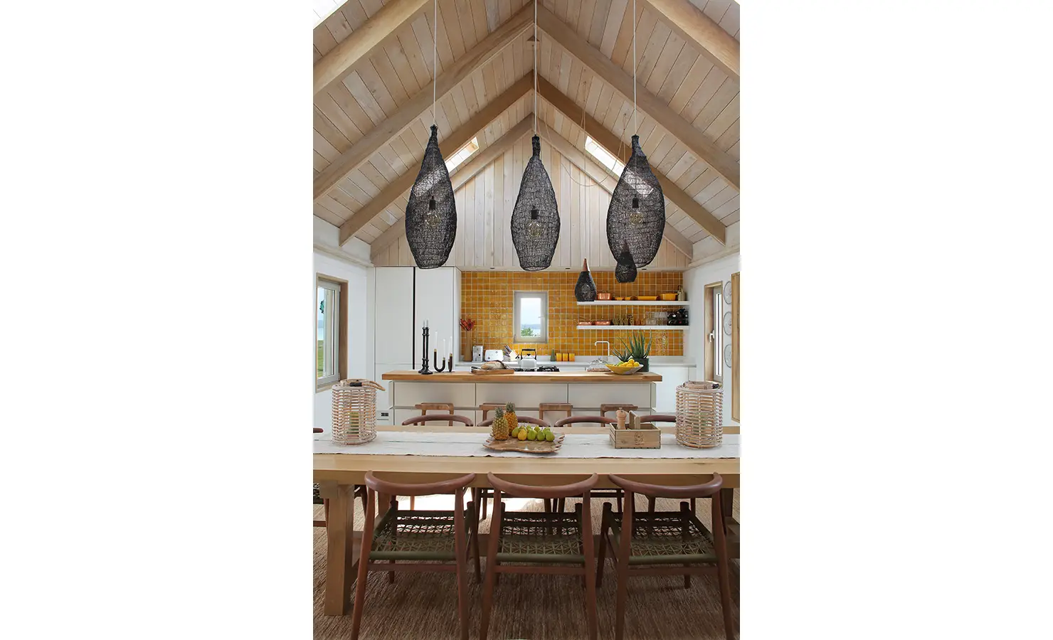 Perfect Hideaways: A beautifully designed dining and kitchen area with a long wooden table, modern chairs, and a striking set of black woven pendant lights, creating a cosy yet sophisticated atmosphere with warm yellow tones. Flamingo, Churchhaven.