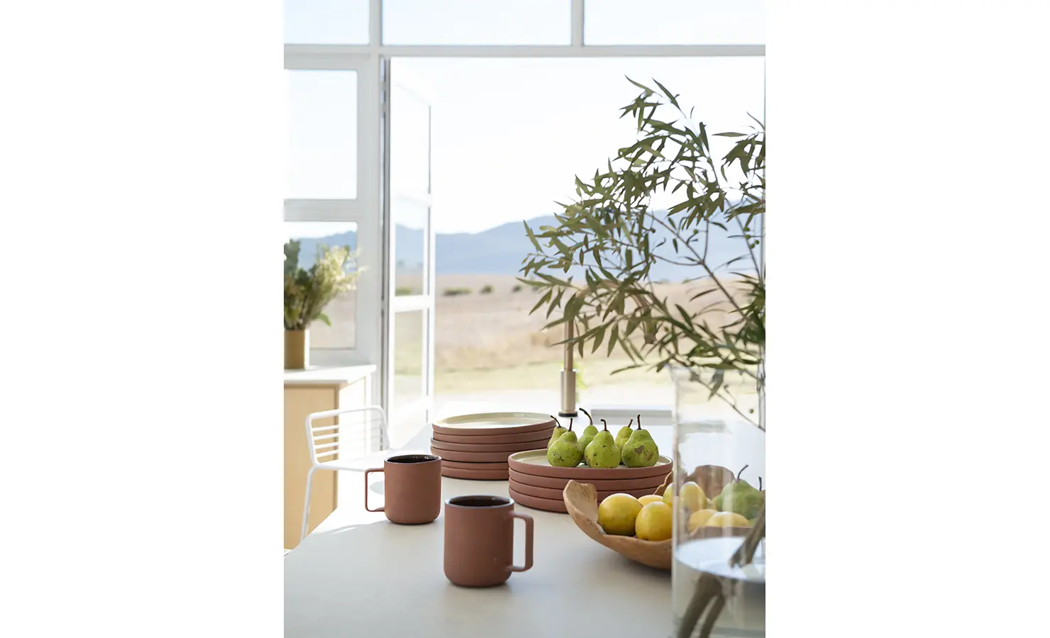 Perfect Hideaways: Breakfast setting with stacked pink plates, mugs, and fresh fruit, overlooking open fields and distant mountains, Thuúla Haven, Hemel-en-Aarde.
