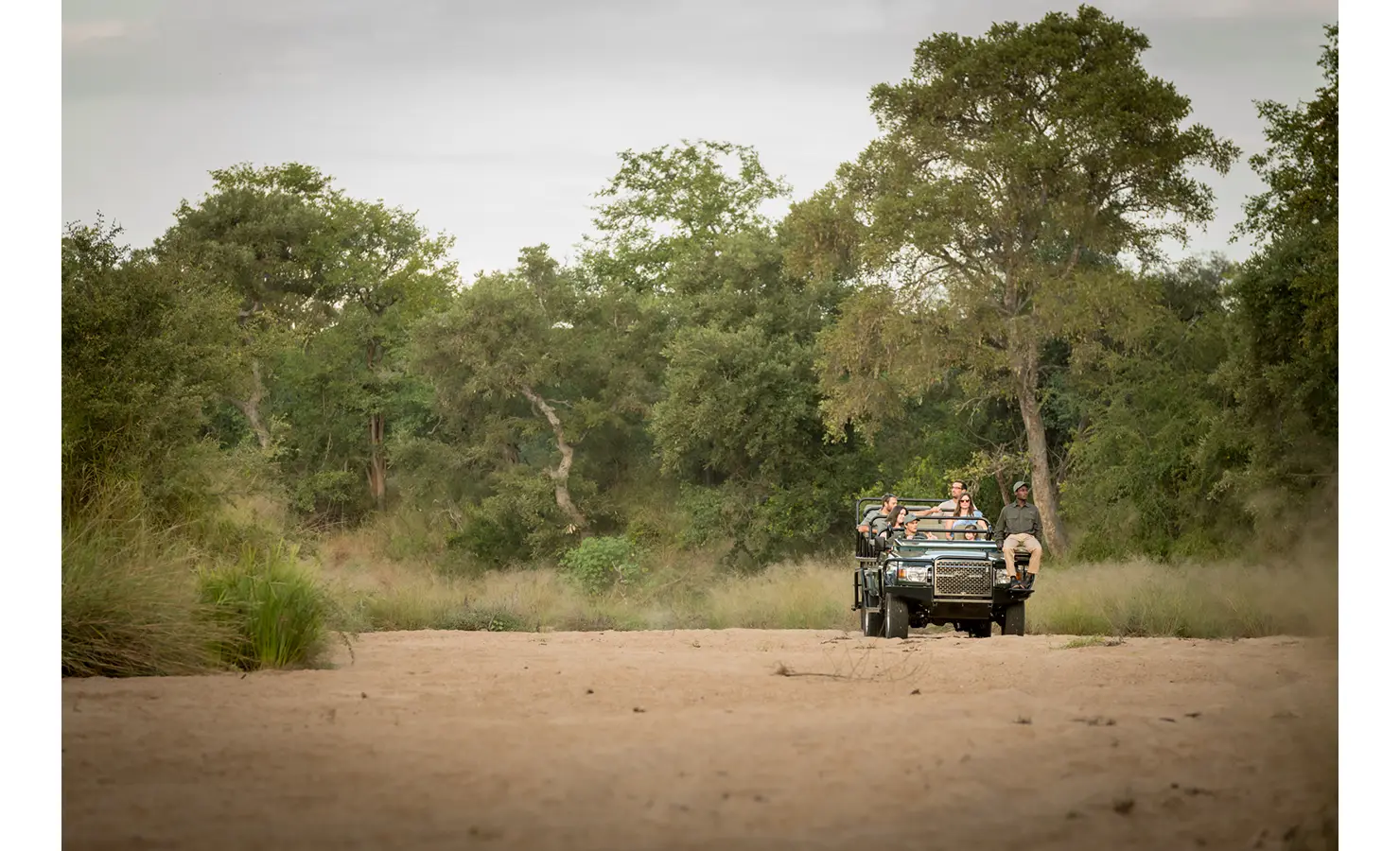 Perfect Hideaways: A safari vehicle driving through a dry riverbed, surrounded by lush trees and bushland, with passengers enjoying the scenic wilderness. Tulela Lodge, Klaserie Private Nature Reserve.