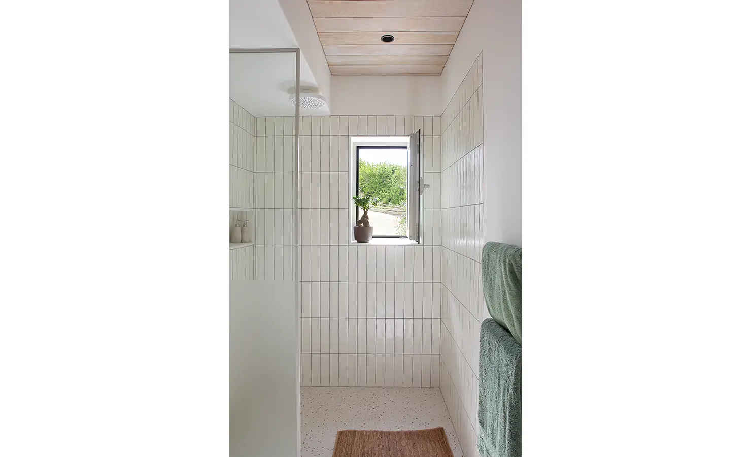 Perfect Hideaways: A minimalist bathroom with soft wooden ceilings and sleek white vertical tiles, featuring a glass-enclosed shower, a small window letting in natural light, and neatly folded green towels. Flamingo, Churchhaven.