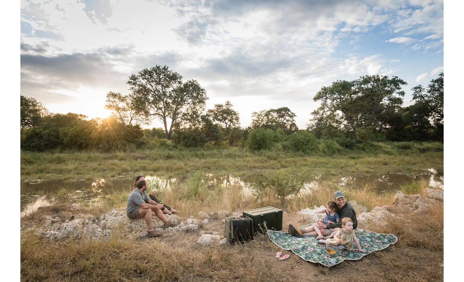 Perfect Hideaways: A tranquil riverside picnic setup with a family enjoying the serene surroundings, as the sun sets over the landscape. Tulela Lodge, Klaserie Private Nature Reserve.