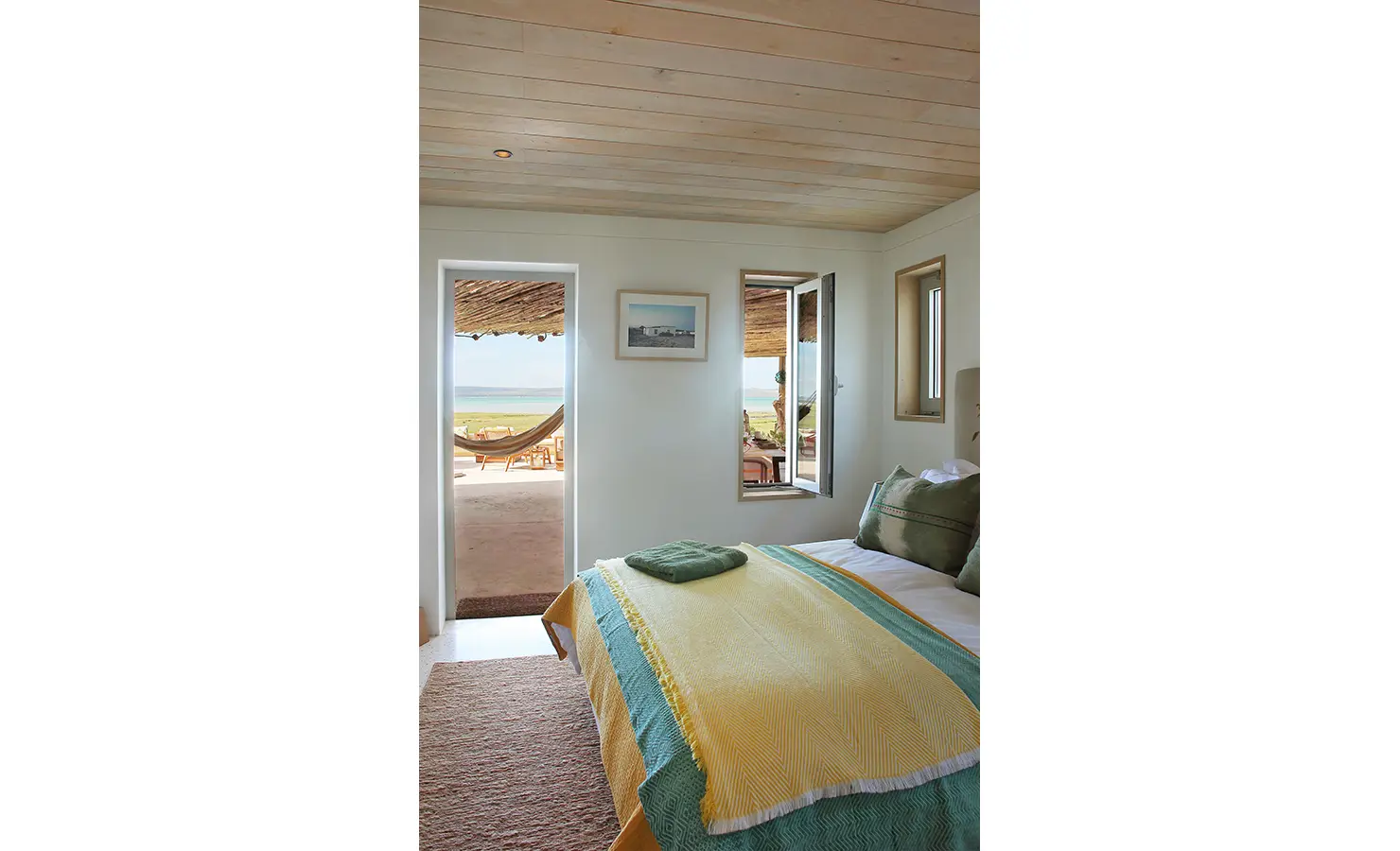 Perfect Hideaways: A serene bedroom with a natural wood ceiling, a soft green and yellow bedspread, and an open doorway leading to a shaded terrace with a hammock and stunning lagoon views. Flamingo, Churchhaven.