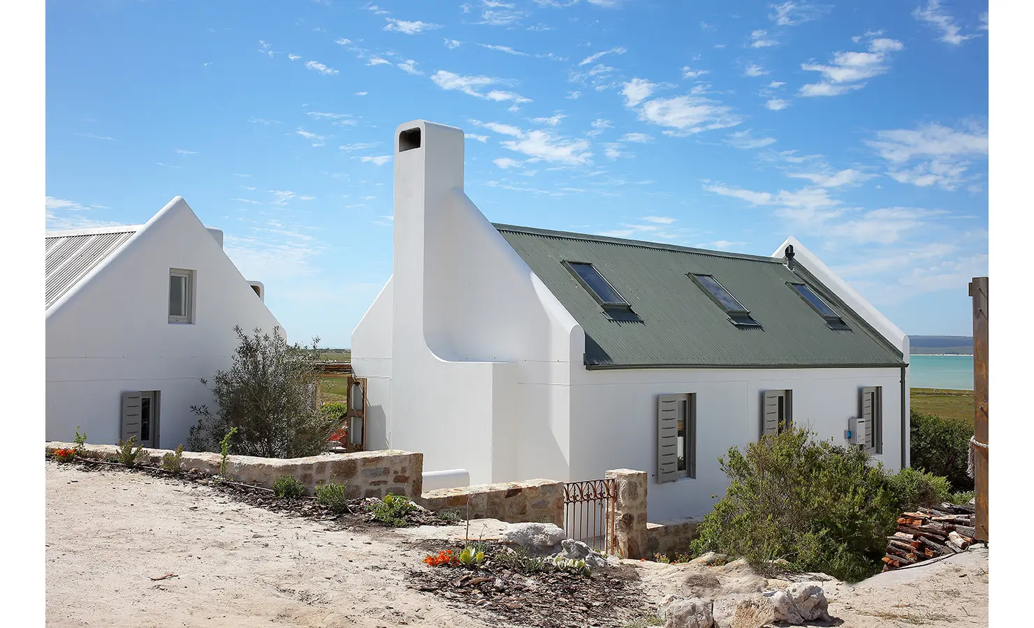 Perfect Hideaways: A charming coastal cottage with whitewashed walls, a traditional chimney, and green window shutters, sitting above a scenic landscape with the lagoon in the background. Flamingo, Churchhaven.