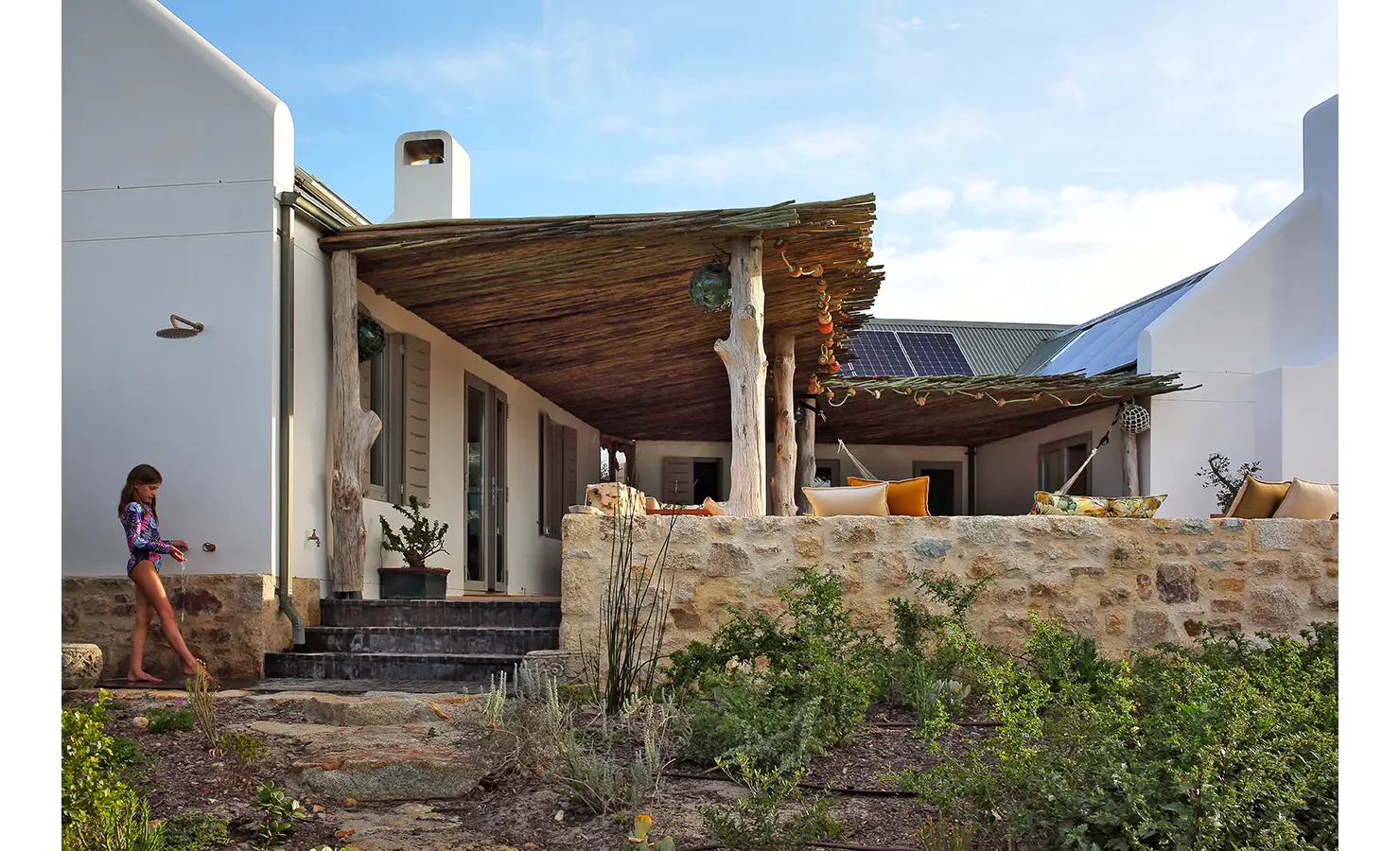 Perfect Hideaways: A rustic outdoor living space framed by thick wooden pillars and a woven reed roof, with a relaxed seating area overlooking a lush indigenous garden and stone pathways. Flamingo, Churchhaven.