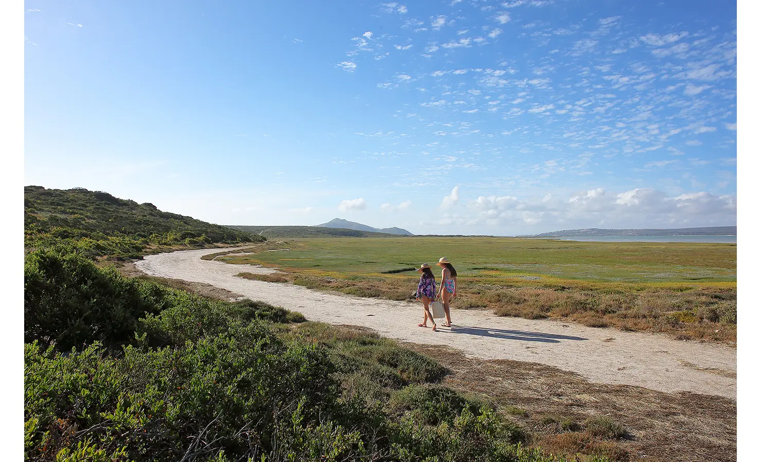 Perfect Hideaways: Two young guests in sun hats and swimsuits walk along a sandy path through the untouched coastal landscape, surrounded by rolling greenery and an expansive blue sky. Flamingo, Churchhaven.