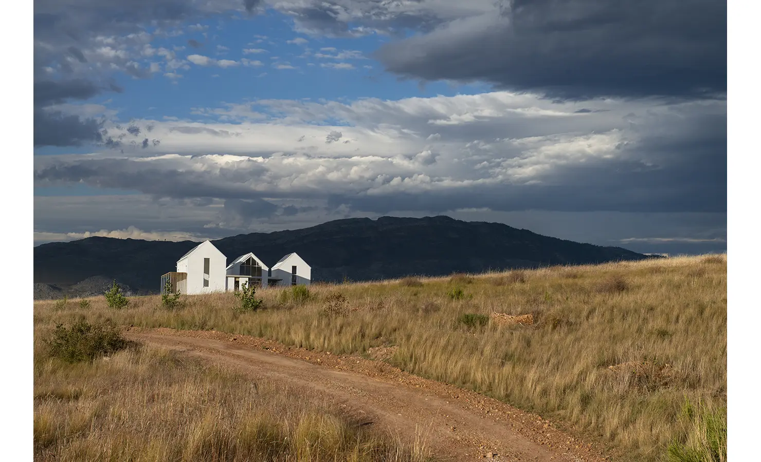 Perfect Hideaways: Contemporary white-gabled home framed by dramatic clouds and mountain backdrop along a rustic dirt track, Thuúla Haven, Hemel-en-Aarde.