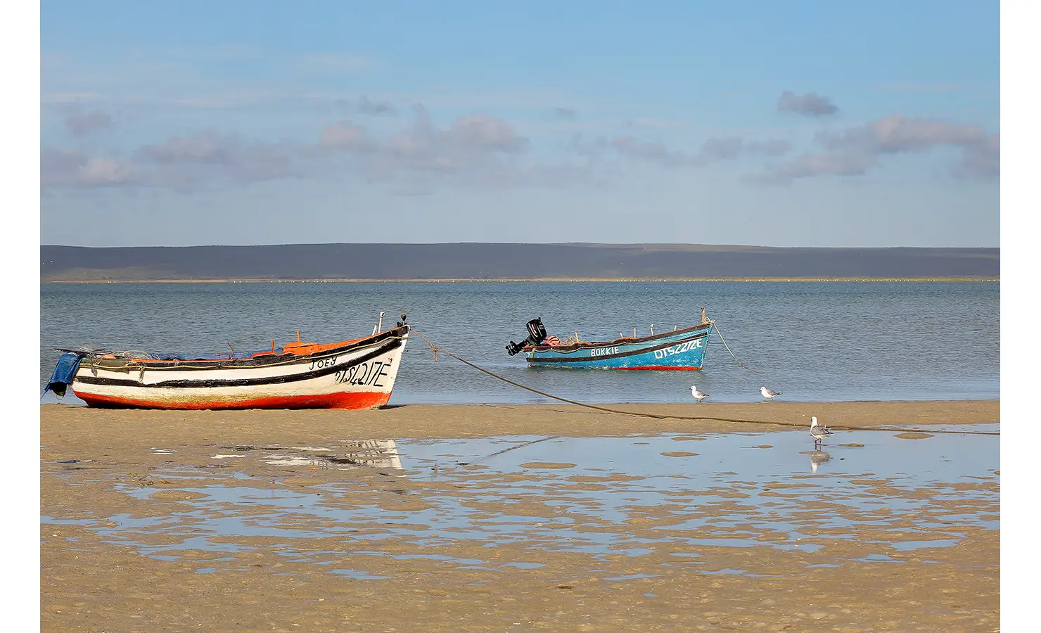 Perfect Hideaways: Two colourful wooden fishing boats rest in the shallow waters of the lagoon, their reflections shimmering on the sand as seagulls wade nearby under a vast blue sky. Flamingo, Churchhaven.