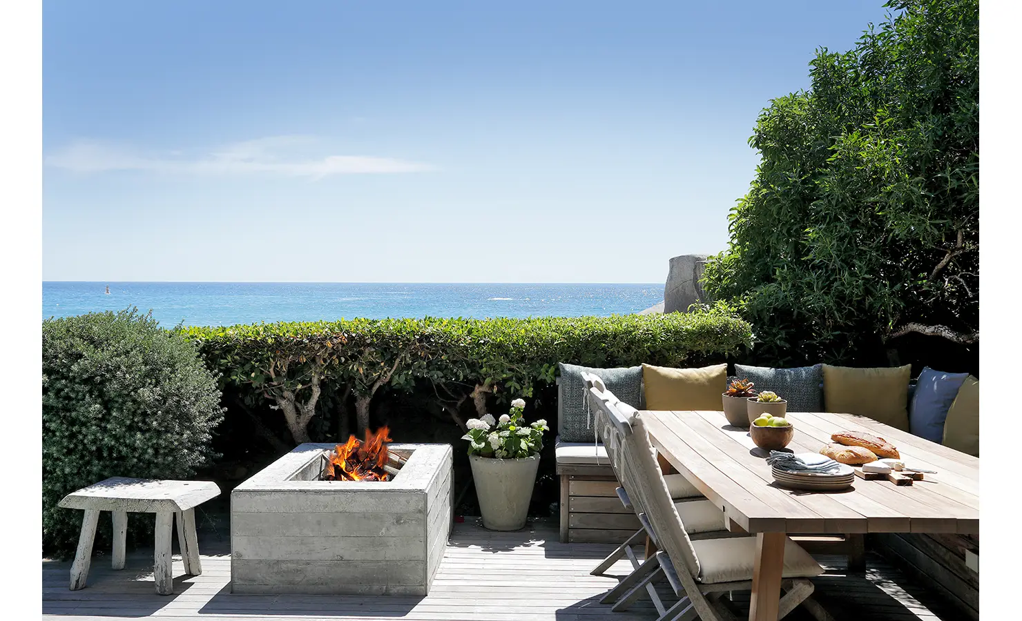 Perfect Hideaways ocean-view outdoor dining terrace with built-in wooden seating, cushioned bench, concrete fire pit, and Atlantic Ocean backdrop Oasis On Glen, Camps Bay