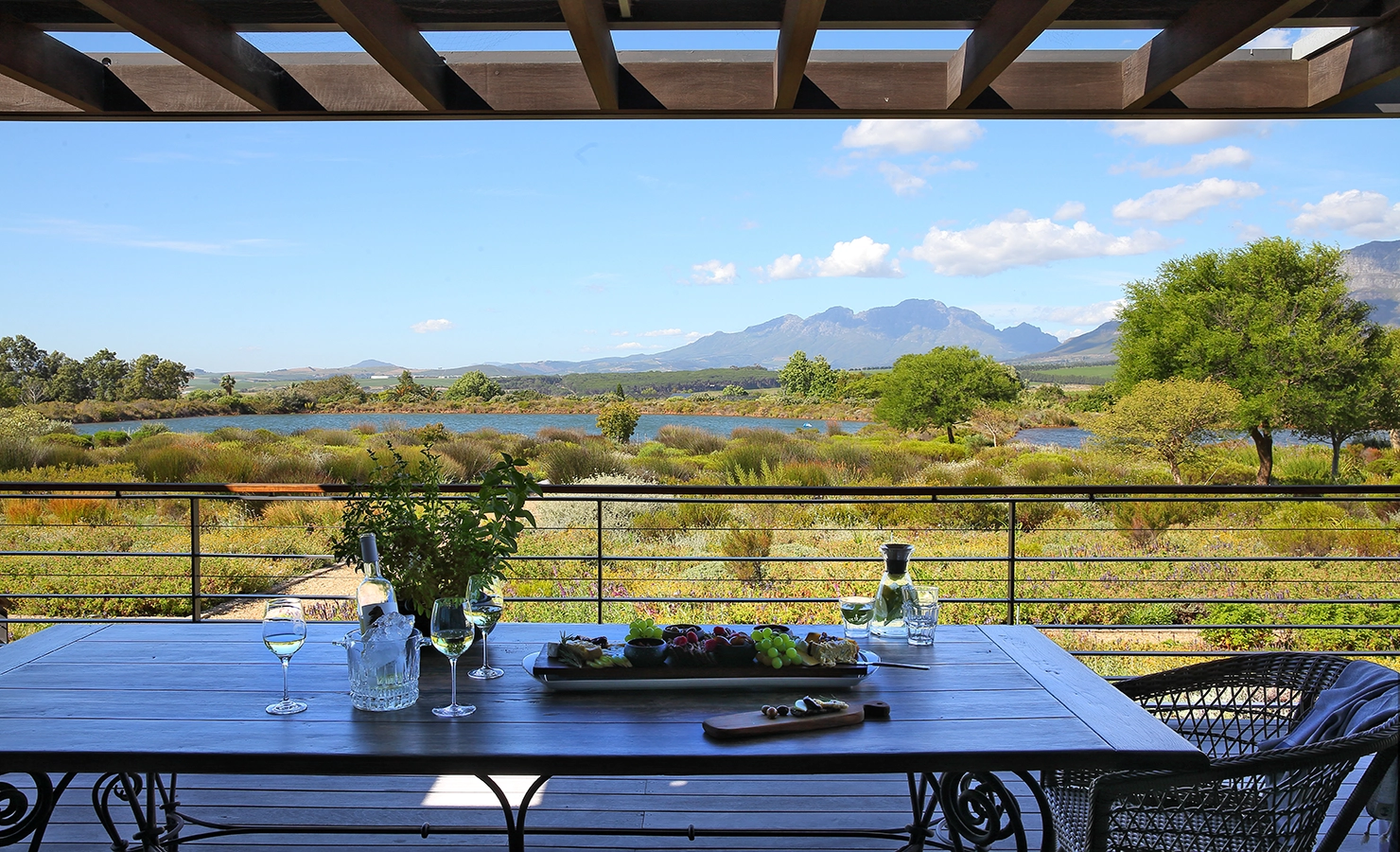 Perfect Hideaways: A scenic outdoor dining setup on a shaded terrace, overlooking a vast landscape of lush greenery, a tranquil lake, and distant mountains under a clear blue sky. A wooden table is set with glasses of chilled white wine, a bottle in an ice bucket, and a tray of fresh fruits and cheeses, inviting a moment of relaxation in the Cape Winelands. Equestrian House, Stellenbosch.