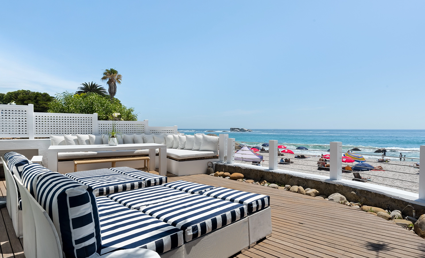 Perfect Hideaways oceanfront terrace with striped navy loungers, white cushioned seating and dining table overlooking Clifton beach with turquoise waters and colourful umbrellas, Clifton Sands, Clifton
