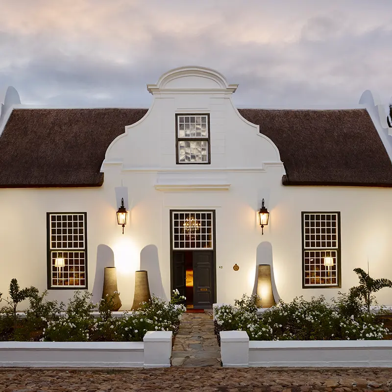Perfect Hideaways elegant Cape Dutch farmhouse with whitewashed façade, thatched roof, symmetrical windows and softly lit entrance framed by manicured garden at dusk Ballotina, Tulbagh